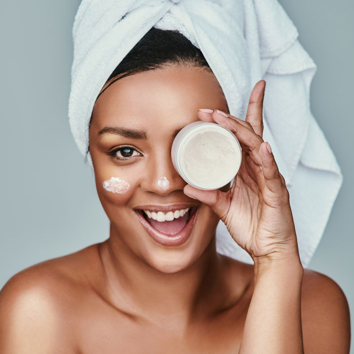 Medium skin toned woman glowing skin, a towel on her head, and beauty product on her face and in her hand with a wide smile