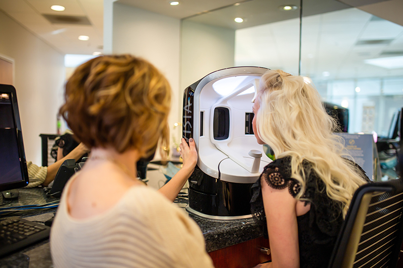 Long haired blond woman sticking face inside of Visia Skin Analysis Device, with assistant walking her through it.