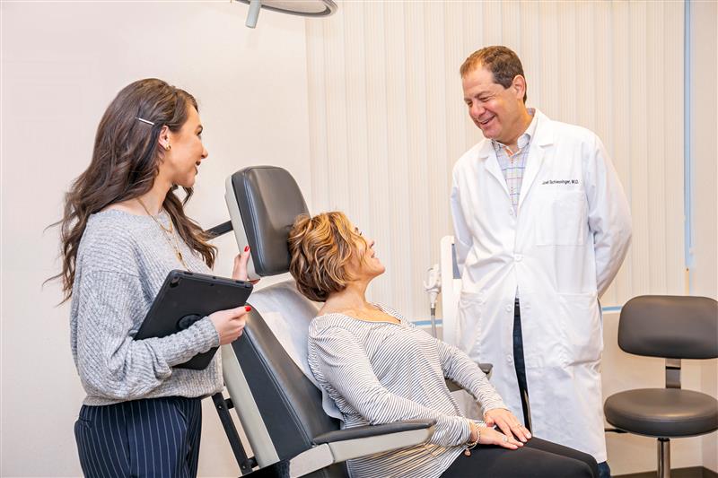 Dr, Schlessinger talking to light skin toned patient in exam chair with his female assistant beside her.