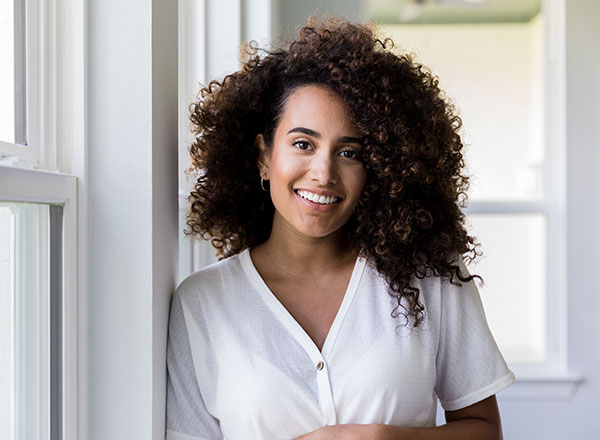 Dark skin toned woman standing by windows smiling at camera